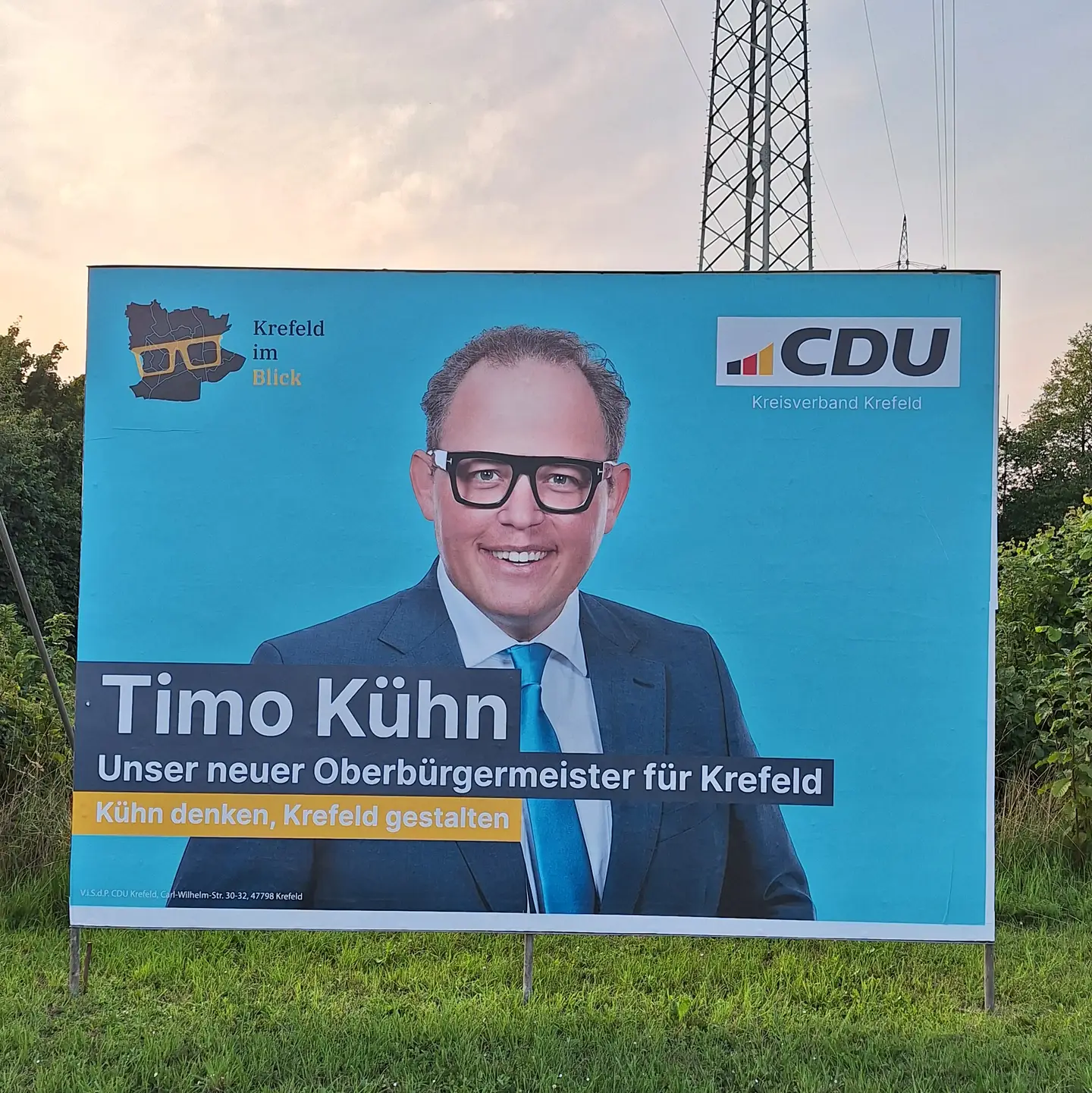 Foto des großen Wahlplakat (Aufsteller) mit unserem Obermeisterkandidat Timo Kühn - im Hintergrund Hochspannungsmast unseres Umspannwerk in Gellep-Stratum